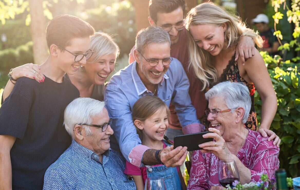 Multi-generational family gathered outdoors laughing together looking at a phone, illustrating bridging generations and maintaining family connections for seniors supported by TheKey
